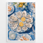 Indlæs billede til gallerivisning Oyster platter art print plakat showing oysters and lemon slices on a blue patterned table with seafood dishes and glassware in a White frame.
