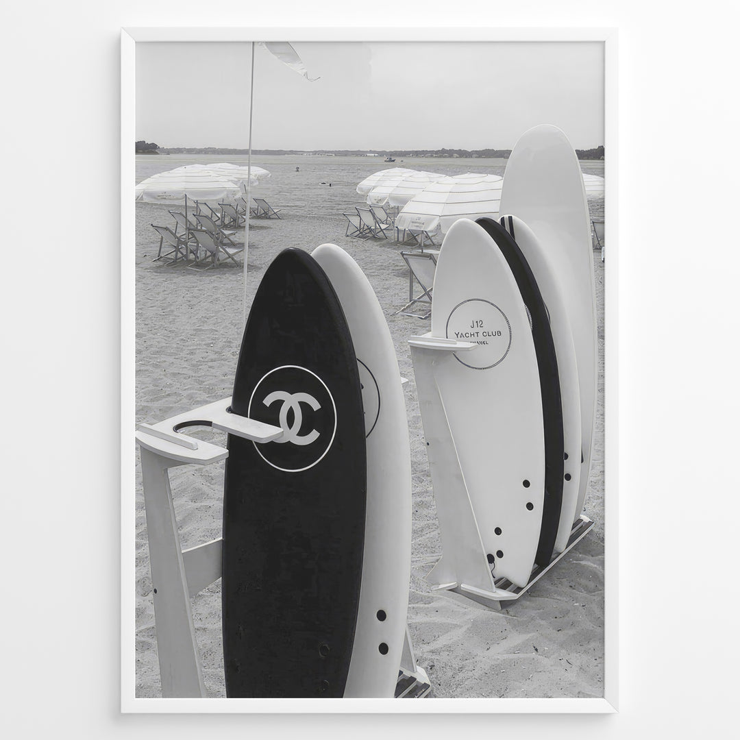 Fashion Plakat | Chanel Surfboards on the Beach – Luxury Meets Coastal Lifestyle in Black and White Plakaty.dk | Black and white photo of Chanel and J12 Yacht Club surfboards on a sandy beach with umbrellas and lounge chairs in the background