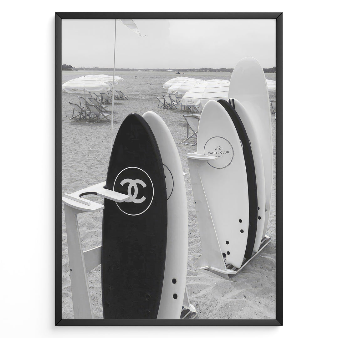 Fashion Plakat | Chanel Surfboards on the Beach – Luxury Meets Coastal Lifestyle in Black and White Plakaty.dk | Black and white photo of Chanel and J12 Yacht Club surfboards on a sandy beach with umbrellas and lounge chairs in the background
