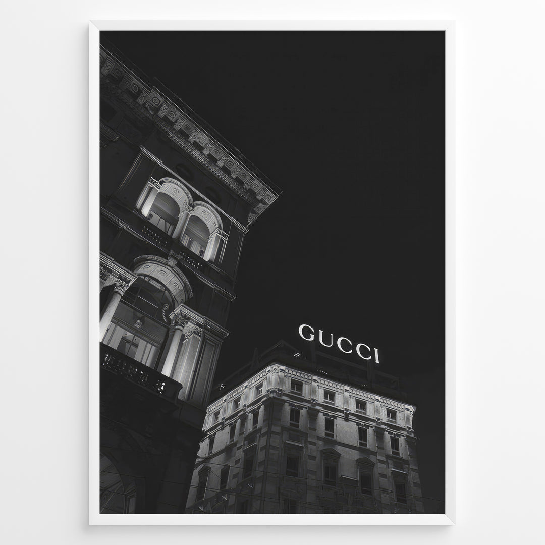 Fashion Plakat | Night View of Gucci Building Facade Plakaty.dk | Black and white nighttime photo of an ornate building with classical columns and arches next to a building displaying a large illuminated "GUCCI" sign against a dark sky.