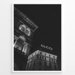 Indlæs billede til gallerivisning Fashion Plakat | Night View of Gucci Building Facade Plakaty.dk | Black and white nighttime photo of an ornate building with classical columns and arches next to a building displaying a large illuminated &quot;GUCCI&quot; sign against a dark sky.

