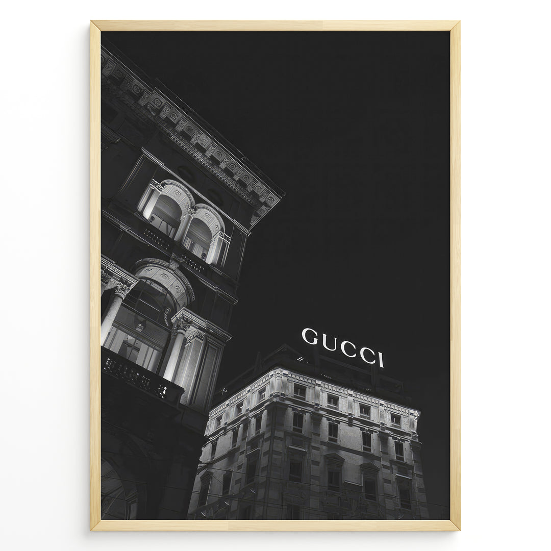 Fashion Plakat | Night View of Gucci Building Facade Plakaty.dk | Black and white nighttime photo of an ornate building with classical columns and arches next to a building displaying a large illuminated "GUCCI" sign against a dark sky.