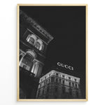 Indlæs billede til gallerivisning Fashion Plakat | Night View of Gucci Building Facade Plakaty.dk | Black and white nighttime photo of an ornate building with classical columns and arches next to a building displaying a large illuminated &quot;GUCCI&quot; sign against a dark sky.
