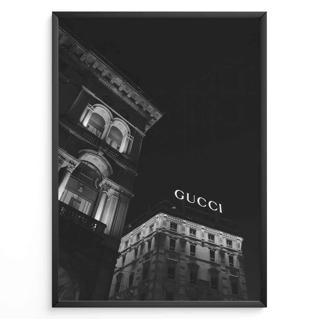 Fashion Plakat | Night View of Gucci Building Facade Plakaty.dk | Black and white nighttime photo of an ornate building with classical columns and arches next to a building displaying a large illuminated "GUCCI" sign against a dark sky.