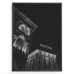 Indlæs billede til gallerivisning Fashion Plakat | Night View of Gucci Building Facade Plakaty.dk | Black and white nighttime photo of an ornate building with classical columns and arches next to a building displaying a large illuminated &quot;GUCCI&quot; sign against a dark sky.
