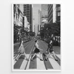 Indlæs billede til gallerivisning Fashion Plakat | Three Women on a Crosswalk in a City Plakaty.dk | Et indrammet sort-hvidt fotografi, der viser tre kvinder, der går fra venstre mod højre på et fodgængerfelt i en by. Kvinden til venstre har en kjole på, kvinden i midten har en top og nederdel, og kvinden til højre har en kort, mørk kjole på og bærer en taske. Høje skyskrabere og et tårn er synlige i baggrunden, med biler parkeret langs gaden.
