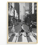 Indlæs billede til gallerivisning Fashion Plakat | Three Women on a Crosswalk in a City Plakaty.dk | Et indrammet sort-hvidt fotografi, der viser tre kvinder, der går fra venstre mod højre på et fodgængerfelt i en by. Kvinden til venstre har en kjole på, kvinden i midten har en top og nederdel, og kvinden til højre har en kort, mørk kjole på og bærer en taske. Høje skyskrabere og et tårn er synlige i baggrunden, med biler parkeret langs gaden.
